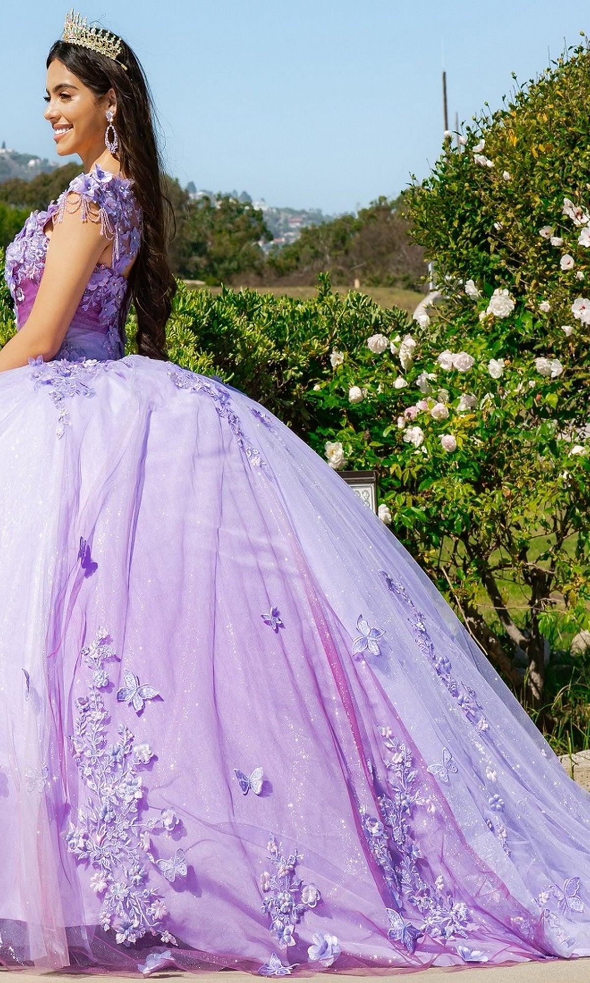 A young woman wearing the Quinceanera Ball Gown 8088J, featuring a tiara, lavender color, 3D floral lace, and butterfly details, stands outdoors near green bushes and white flowers.